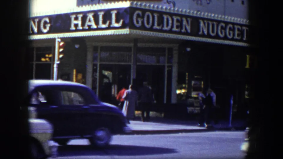 A dark sedan crosses the foreground as pedestrians stand near the Golden Nugget frontage in 1954 Las Vegas.