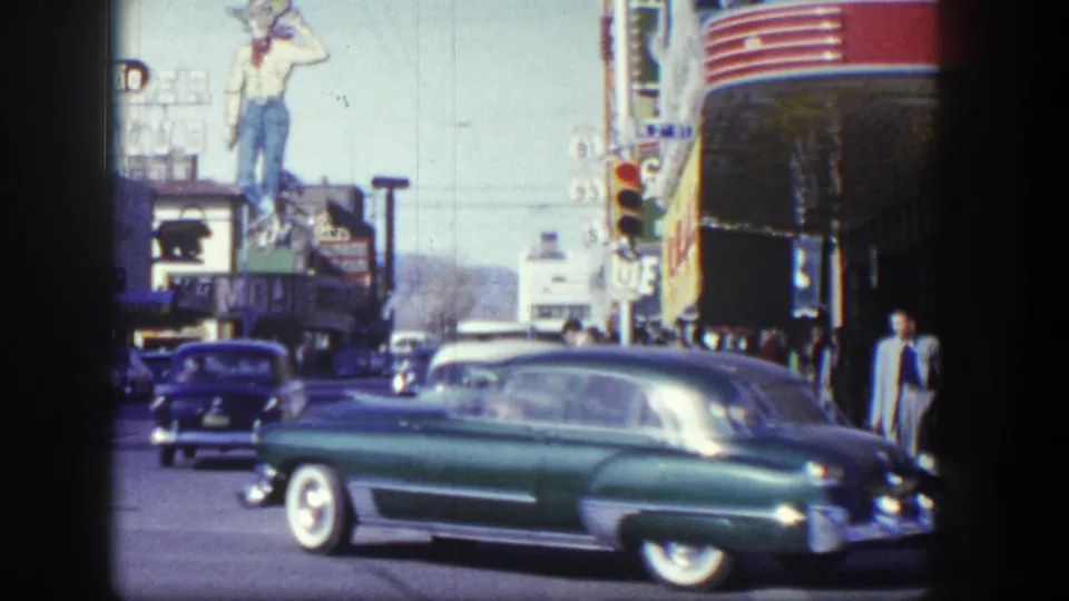 Downtown Las Vegas street scene with a green mid-century car passing traffic lights and large commercial signs.