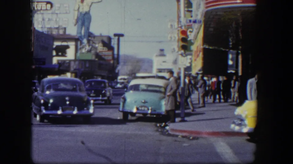 1954 Las Vegas street scene with mid-century cars, pedestrians near the curb, traffic signals, and large commercial neon signs.