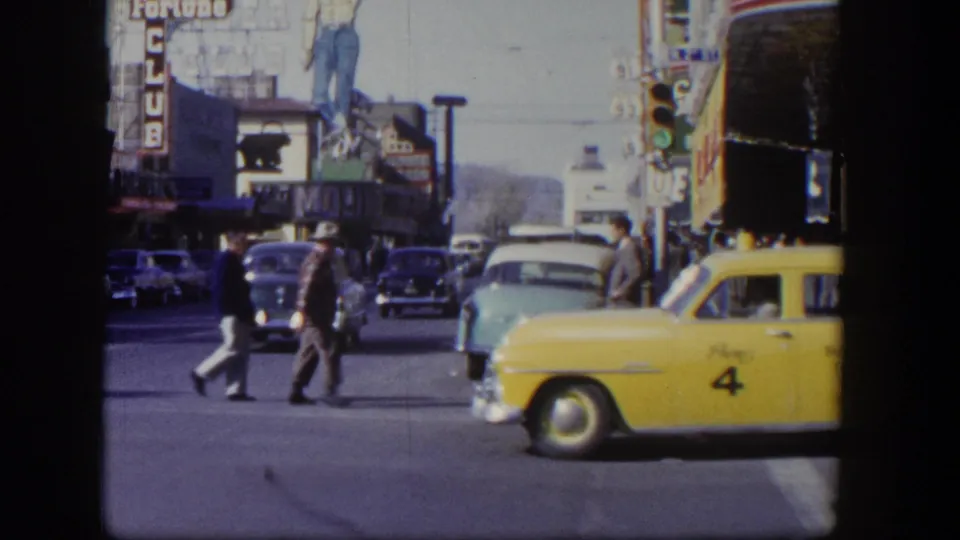 A yellow taxi marked with number 4 crosses a 1954 Las Vegas intersection as pedestrians and older cars appear behind it.
