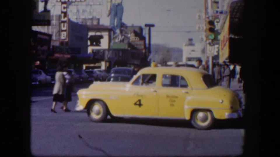 The yellow taxi moves across the foreground of a 1954 Las Vegas street scene with pedestrians, parked cars, and neon signage beyond.
