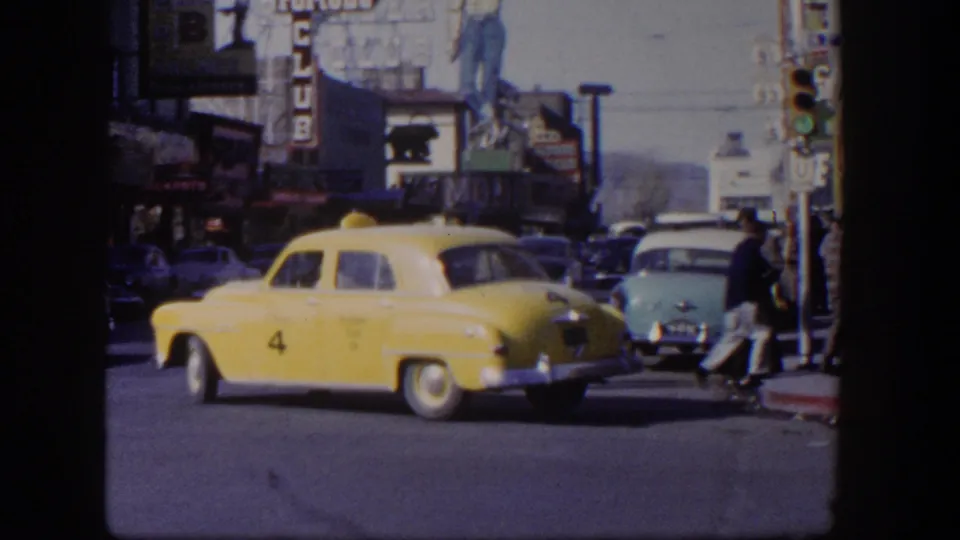 A yellow taxi exits across a Las Vegas intersection in 1954, with a pale blue sedan, pedestrians, and commercial signage visible behind it.