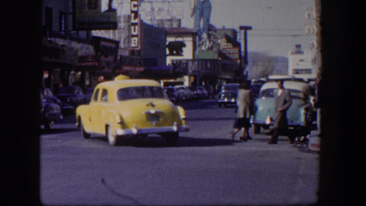 Yellow taxi driving on a city street with classic cars, pedestrians, and storefront signs.