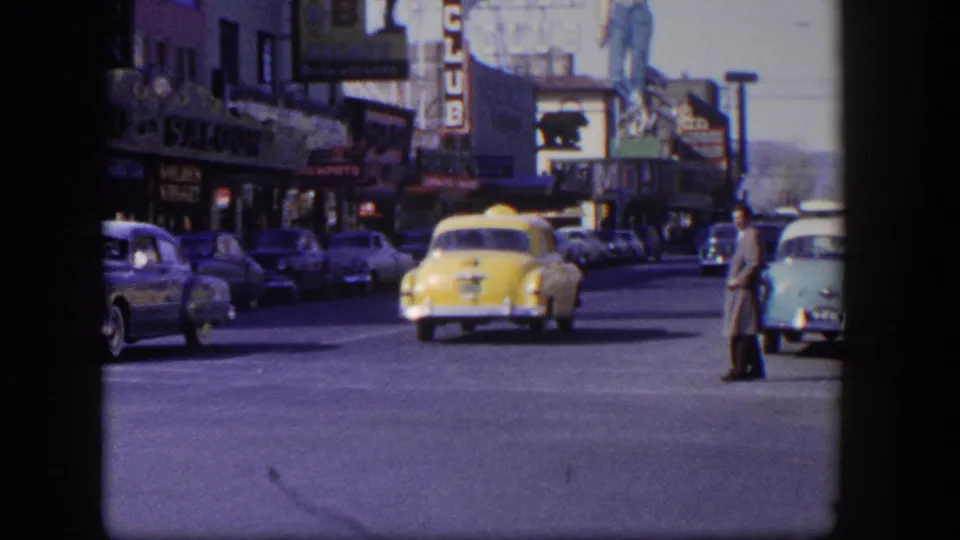 Street-level Las Vegas traffic scene with a yellow taxi, parked automobiles, a standing pedestrian, and visible club signage.