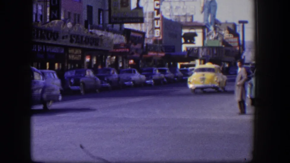1954 Las Vegas street scene with the yellow taxi moving away beneath storefront signs and rows of parked classic cars.