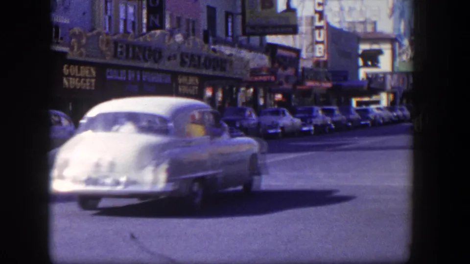 A light-colored 1950s car passes close to the camera on a Las Vegas street with parked cars and business signage behind it.