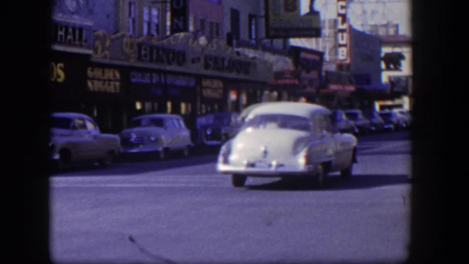 1954 Las Vegas street view with a white sedan moving through traffic below gambling hall, bingo saloon, and food-market signs.