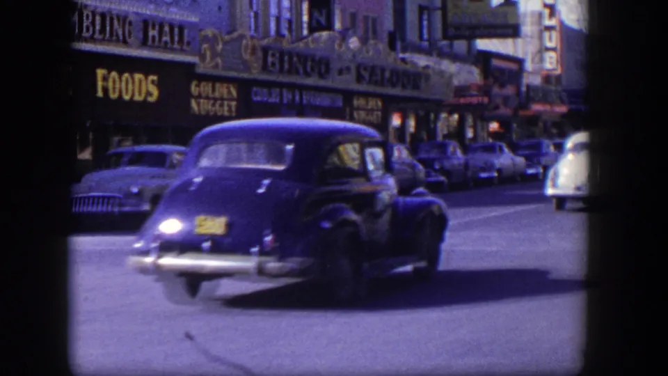 Dark blue mid-century coupe moves across a Las Vegas street in front of gambling hall, bingo, and market signage.