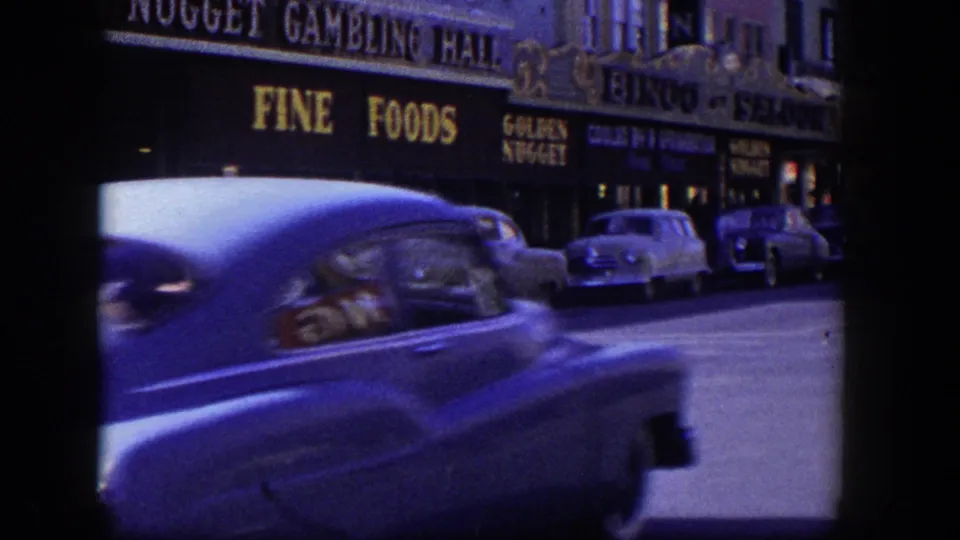 A blue mid-century car passes close in the foreground while Las Vegas food and gambling signage remains visible behind it.