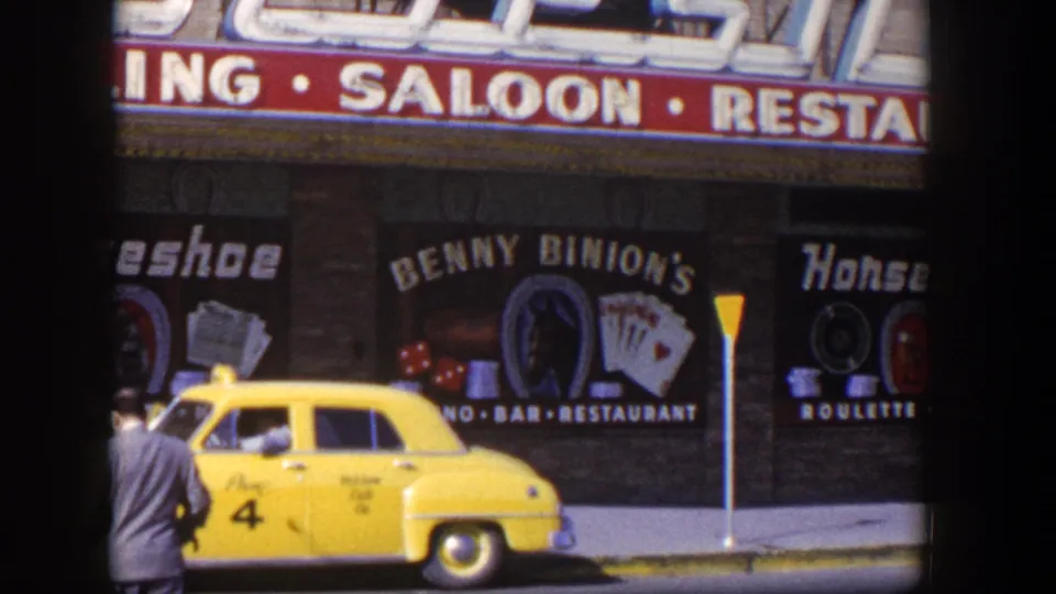 1954 Las Vegas frame showing a yellow taxi and partial pedestrian in front of Benny Binion's Horseshoe casino-front signage.