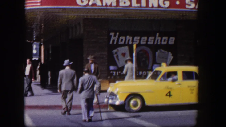 1954 Las Vegas street crossing with men in suits walking toward a curb near a yellow taxi and Horseshoe gambling signage.