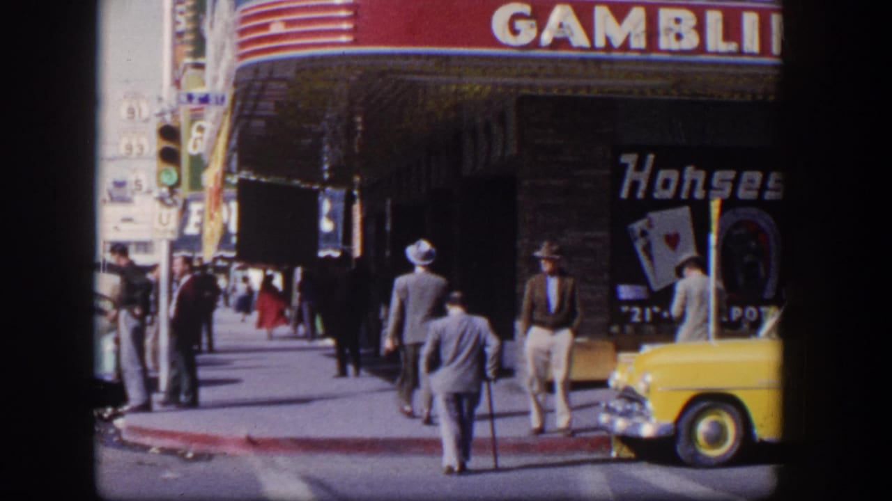 Men in suits and hats walk along a Las Vegas sidewalk beside gambling hall signage and a yellow car.