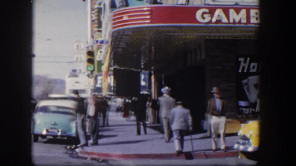 1954 Las Vegas street corner showing pedestrians, a teal car, a yellow car, and storefront gambling signage.