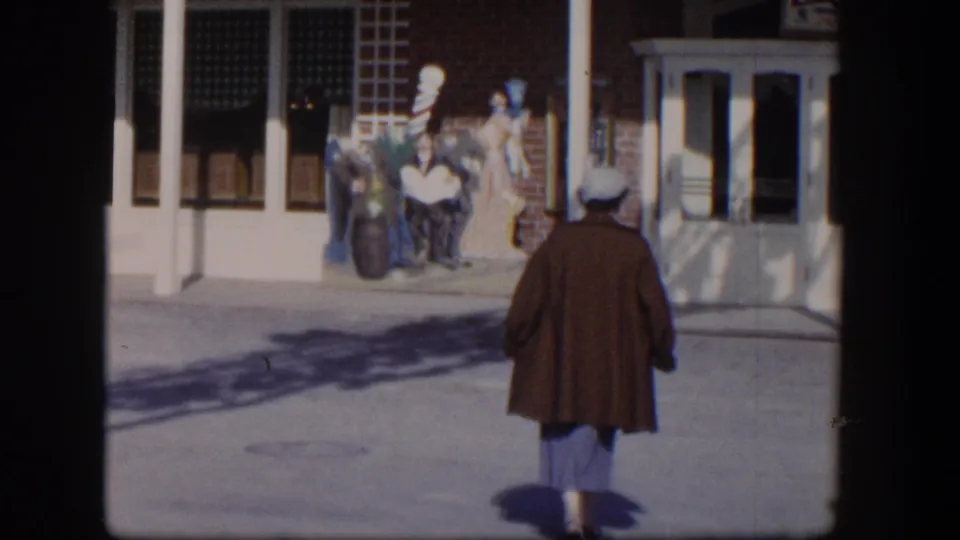 Seen from behind, the woman walks away along the sunlit sidewalk toward storefront windows and a mannequin display in 1954 Las Vegas.