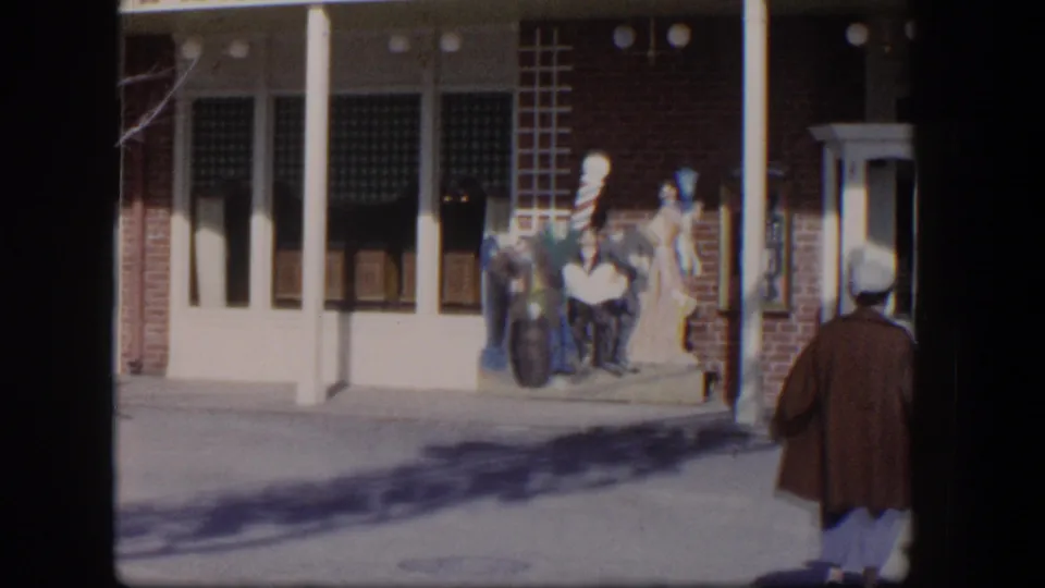 The woman moves toward the edge of the frame as the storefront display, barber pole, brick wall, and sidewalk remain visible.