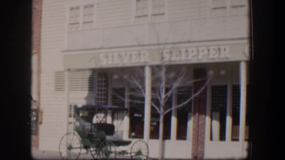 1954 Las Vegas frame showing the Silver Slipper sign, pale building front, bare tree, and parked green carriage.