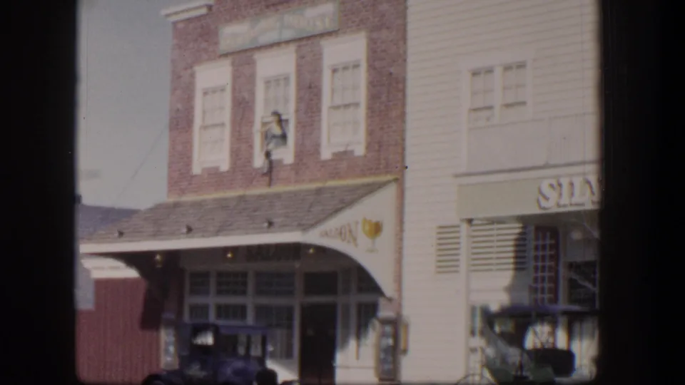 1954 Las Vegas home-movie frame with saloon signage, brick upper facade, parked early automobile, carriage, and partial Silver Slipper storefront.