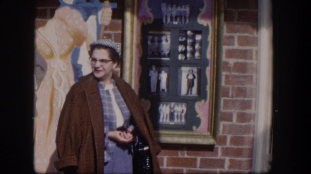 Woman in a brown coat stands by a brick wall and framed display beside painted figures.