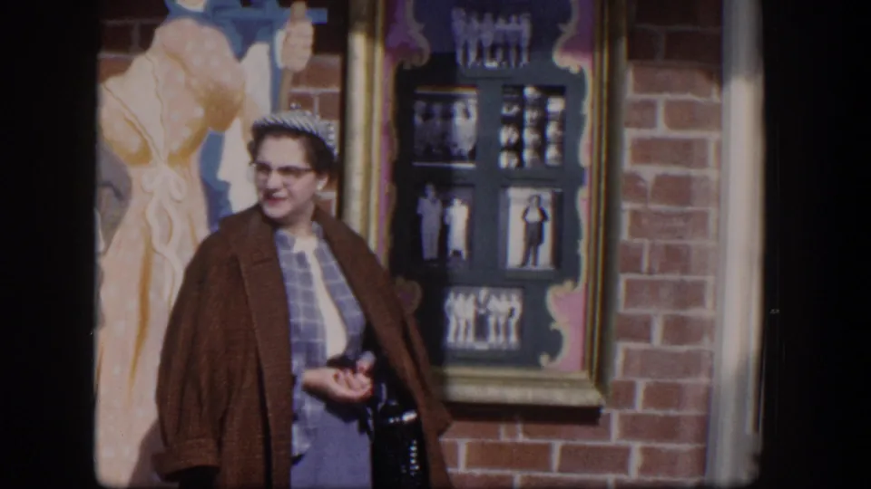 In 1954 Las Vegas footage, a woman in glasses stands near a brick facade, framed display, and colorful barber-themed mural art.
