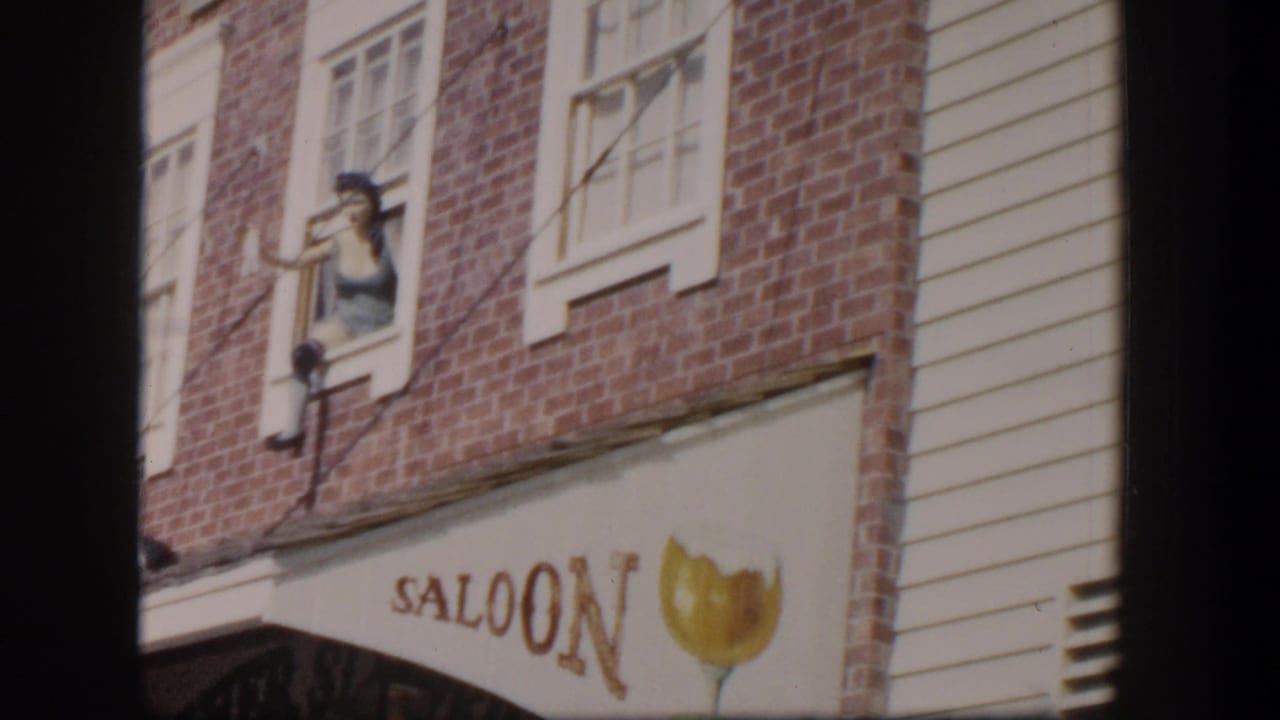 Brick saloon facade with upper windows, painted SALOON sign, and a woman leaning from a window.