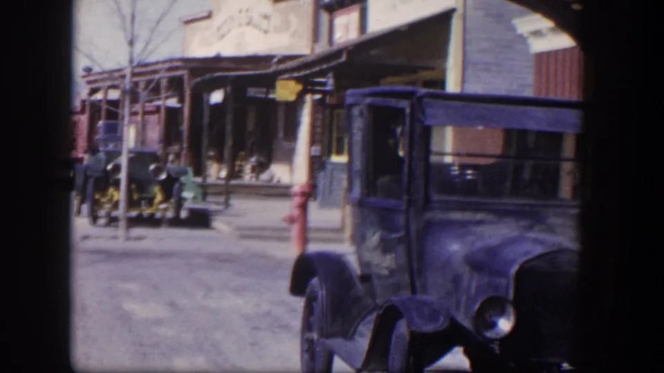 1954 Las Vegas street scene showing a dark truck near the camera, older storefronts, and a second vintage car in the background.