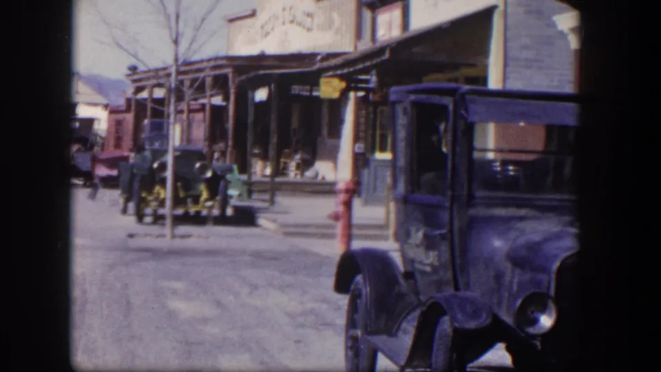1954 Las Vegas home-movie frame with older cars near storefronts, a red hydrant, and a dark parked truck partly filling the right side.