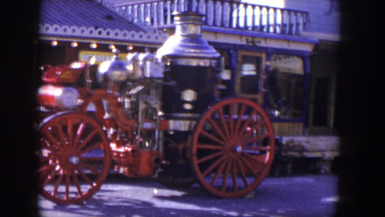 Steam fire engine with red wheels parked before a storefront with balcony railing.