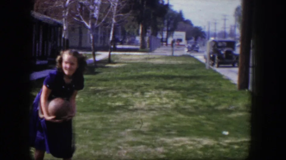 A young woman holds a ball on a lawn beside the street in the same 1954 Las Vegas home-movie sequence.