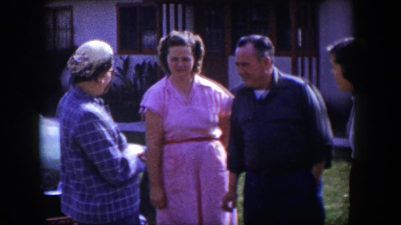 1954 Las Vegas home-movie frame of adults conversing in a yard near a rounded light-colored car and house facade.