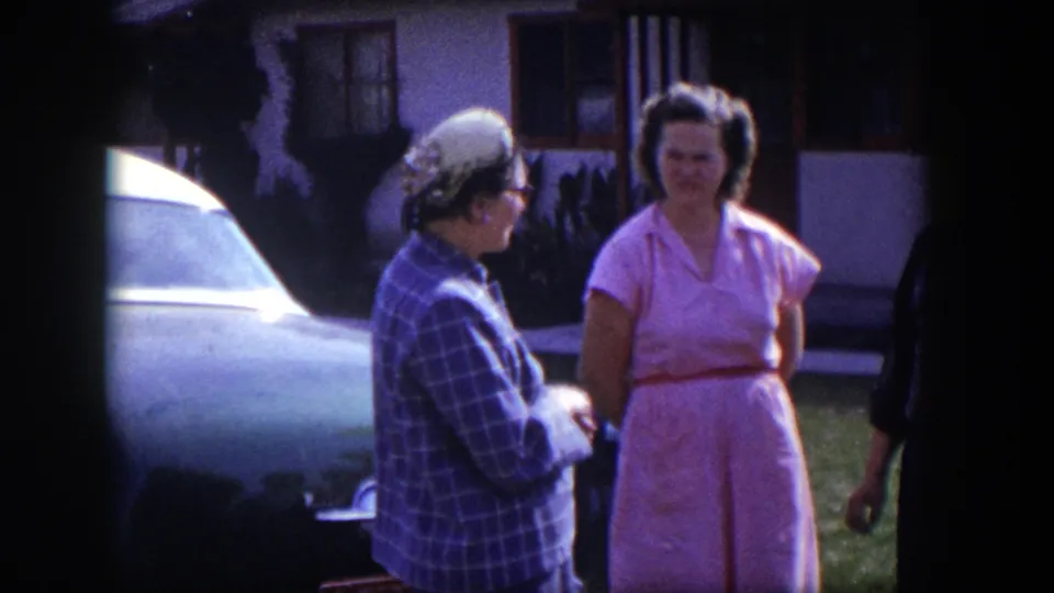 1954 Las Vegas frame showing two women in a yard beside a light-colored car, with part of another person at the edge.