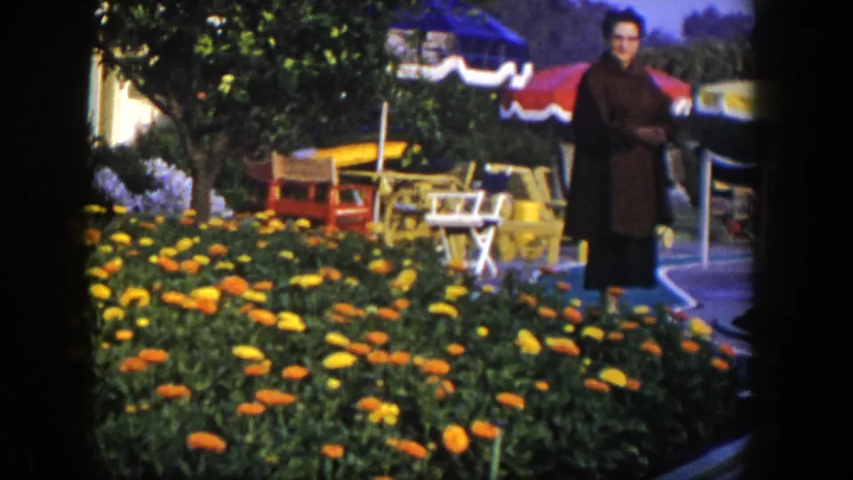 1954 Las Vegas frame showing a woman in a dark coat and glasses near orange and yellow flowers, chairs, and umbrellas.