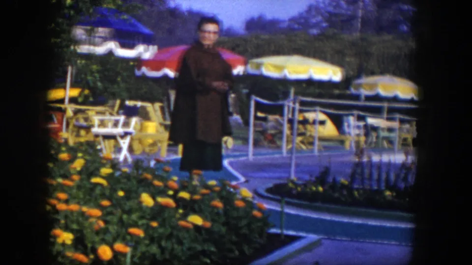 1954 Las Vegas frame with a woman on a paved walkway near vivid flower beds, umbrellas, patio chairs, and poolside paving.