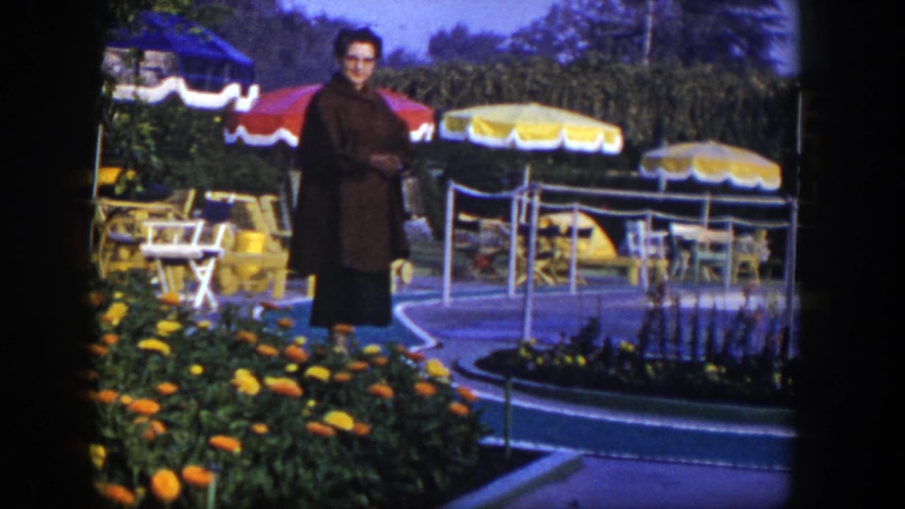 In 1954 Las Vegas footage, a woman in glasses stands near marigolds, patio chairs, and striped umbrellas in an outdoor resort setting.
