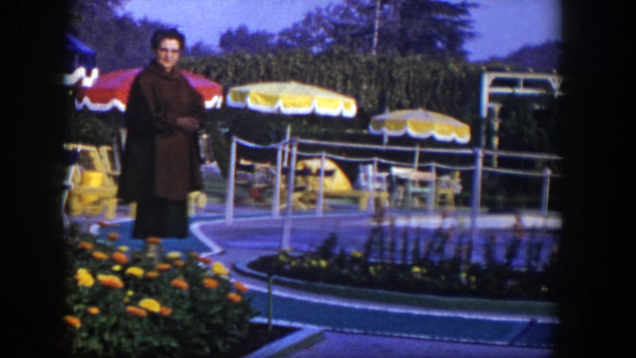 Woman standing near a landscaped pool area with yellow umbrellas and chairs.