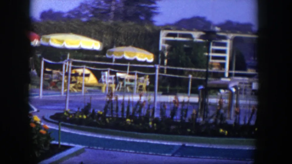 The camera pans across the 1954 Las Vegas resort area, showing umbrellas, patio seating, flower beds, railings, and low structures beyond.