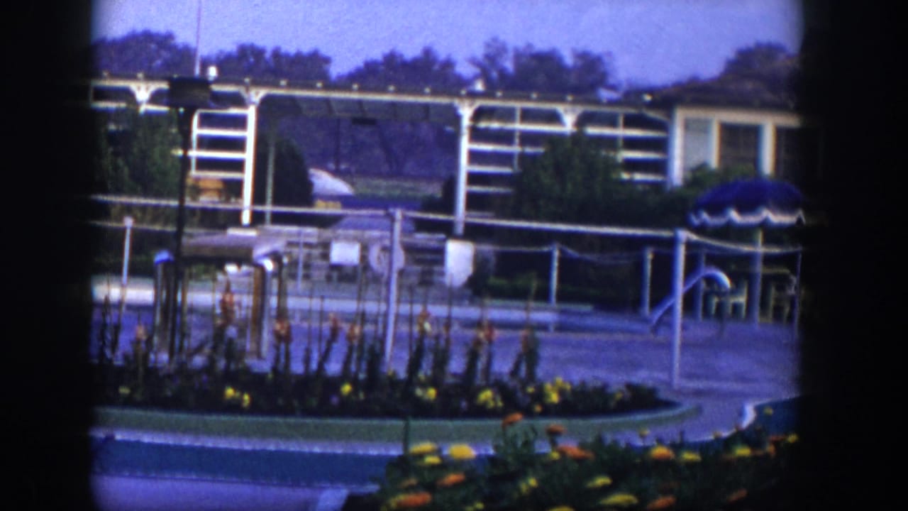 Outdoor seating area with white framework, railings, shrubs, flower beds, and a blue umbrella at right.