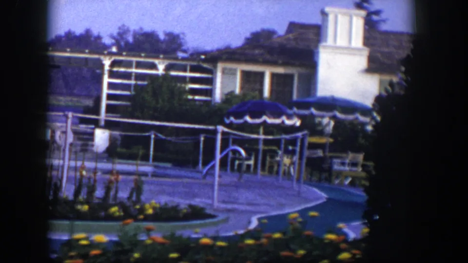 1954 Las Vegas frame showing a curving paved path, blue patio umbrella, chairs, white metal framework, flower beds, and a building exterior.