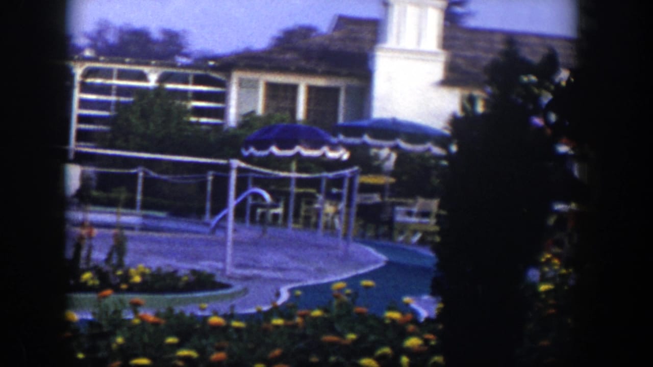 Backyard pool area with flowers, patio umbrellas, chairs, and a white house behind railings.