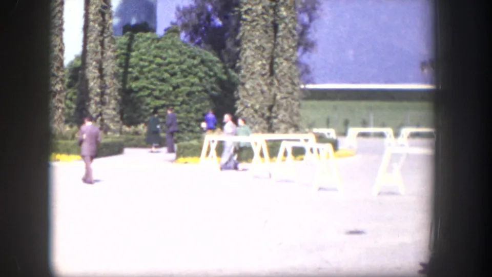 1954 Las Vegas home-movie frame with formally dressed guests near white sawhorse barriers and tall palms outside a light-colored building.