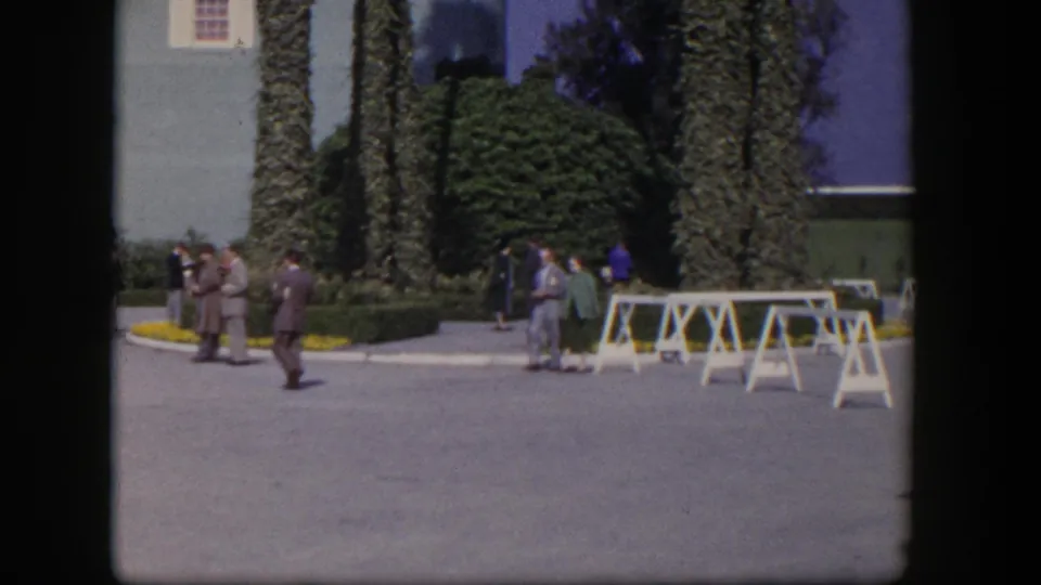 1954 Las Vegas home-movie frame showing adults in suits and dresses moving through a paved outdoor gathering area with palms and white barriers.