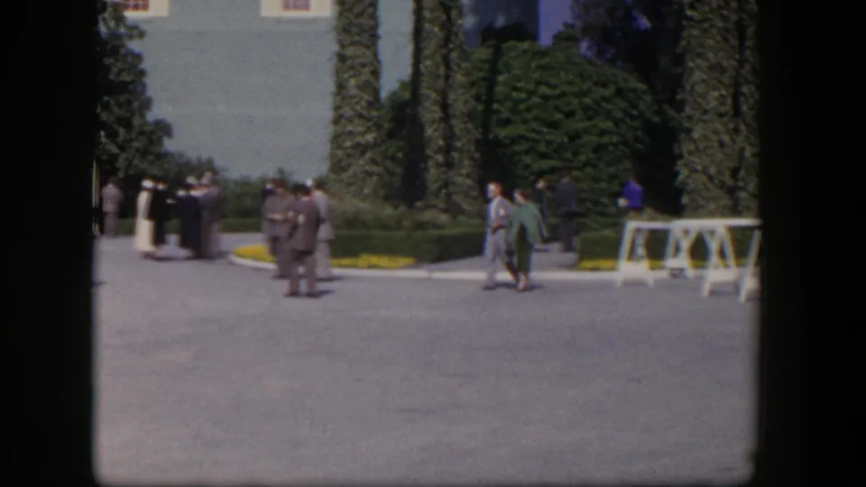 1954 Las Vegas home-movie frame with small groups of formal guests gathered near a landscaped entrance, palms, and white sawhorse barriers.