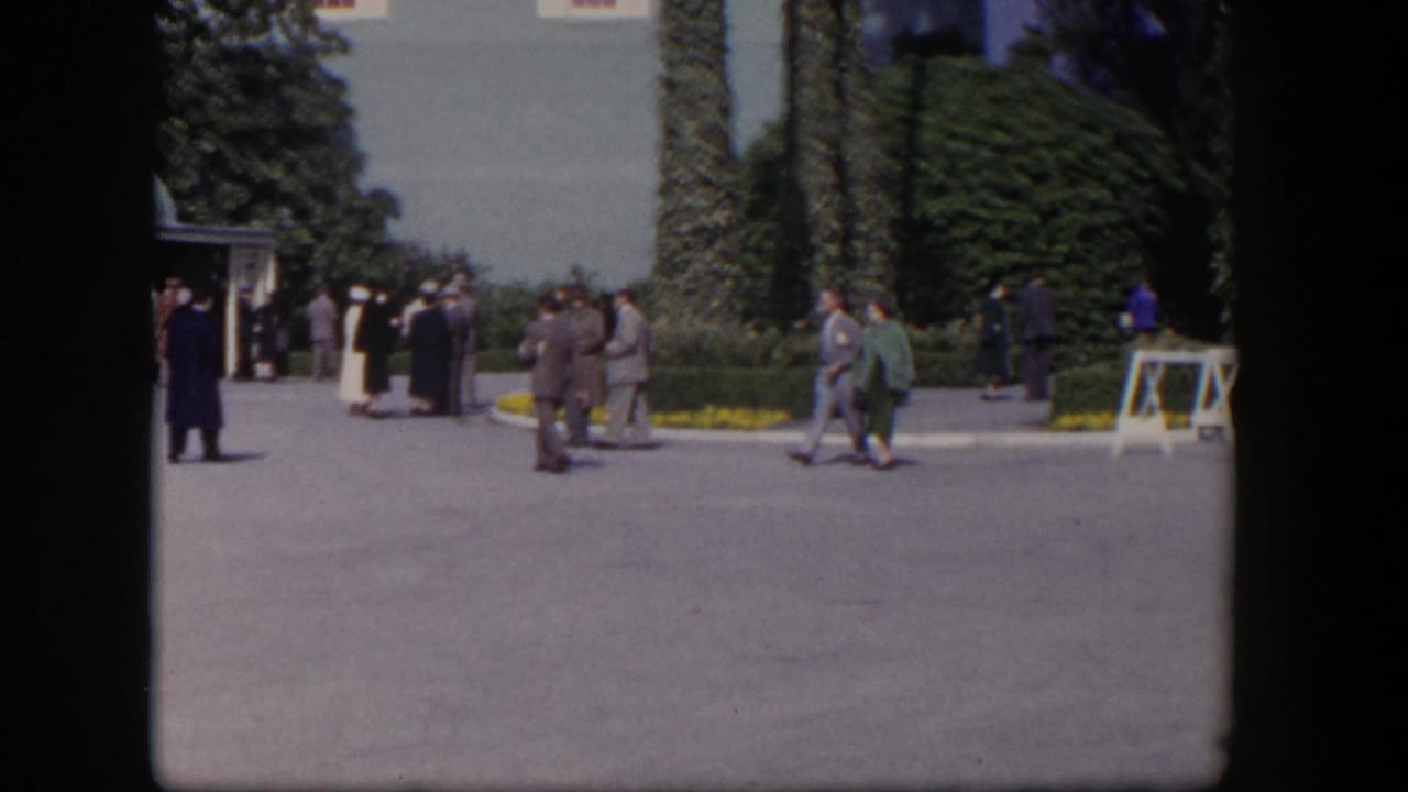 1954 Las Vegas home-movie frame showing adults in suits and dresses crossing a landscaped courtyard near an ivy-covered building.
