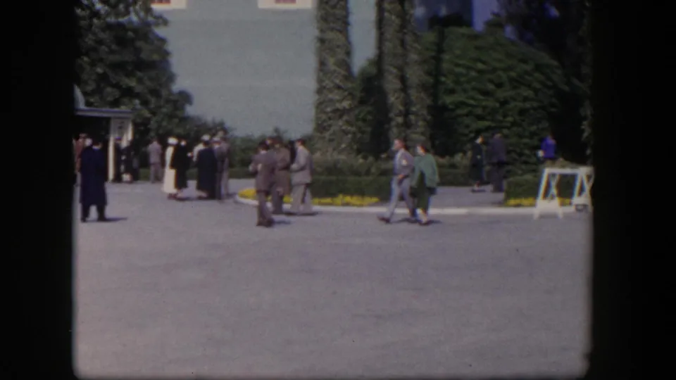 1954 Las Vegas home-movie frame showing adults in suits and dresses crossing a landscaped courtyard near an ivy-covered building.