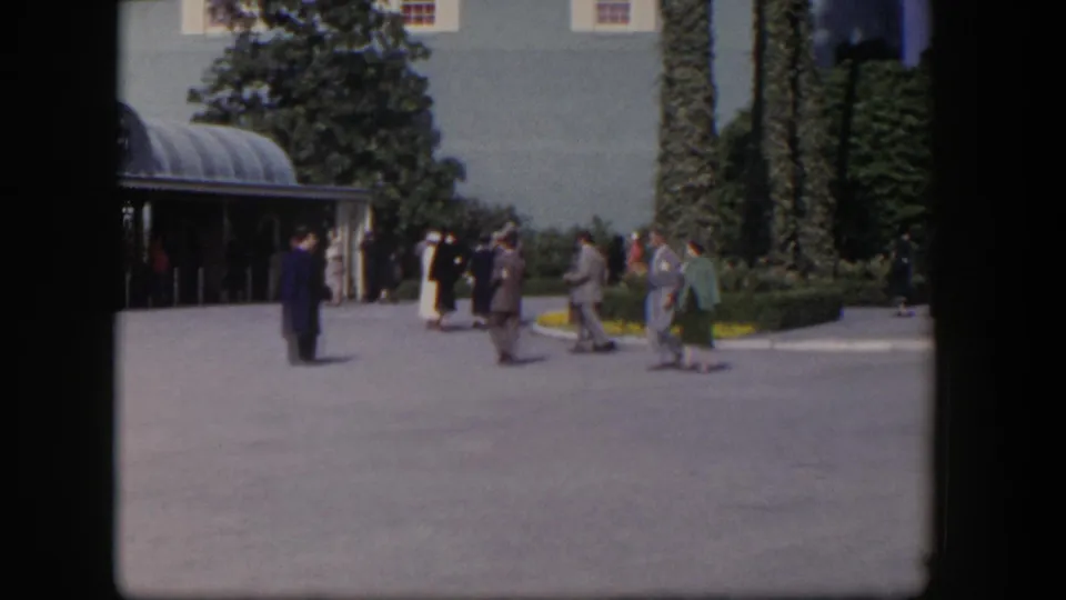 1954 Las Vegas frame with dressed adults walking past a curved glass canopy and landscaped building exterior.