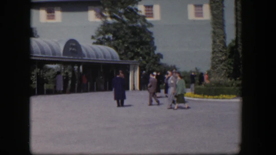 1954 Las Vegas 8mm frame showing visitors in dress clothes moving through a sunny courtyard near an entrance canopy.