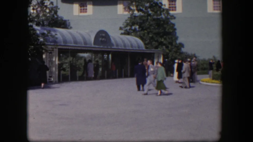 1954 Las Vegas home-movie frame with adults in suits and dresses crossing a plaza by a curved entrance canopy.