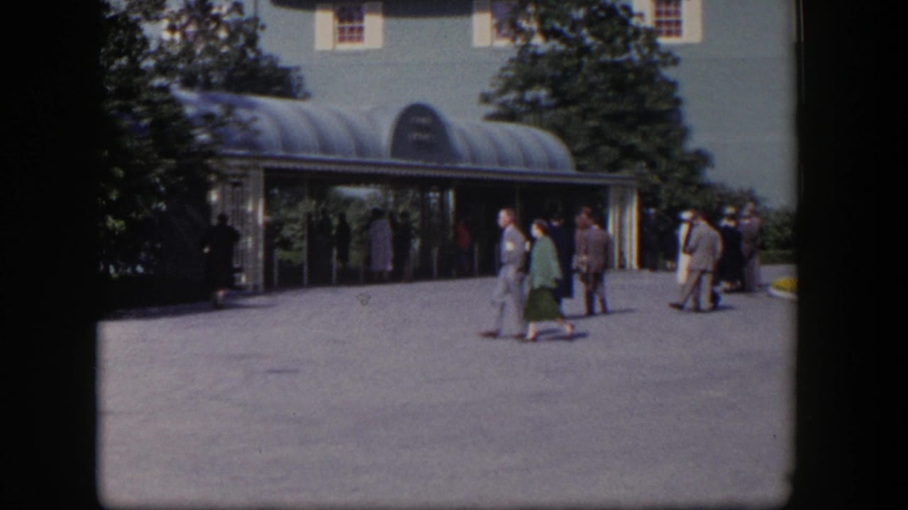 Adults walk across a paved courtyard toward a glass-canopied building entrance.