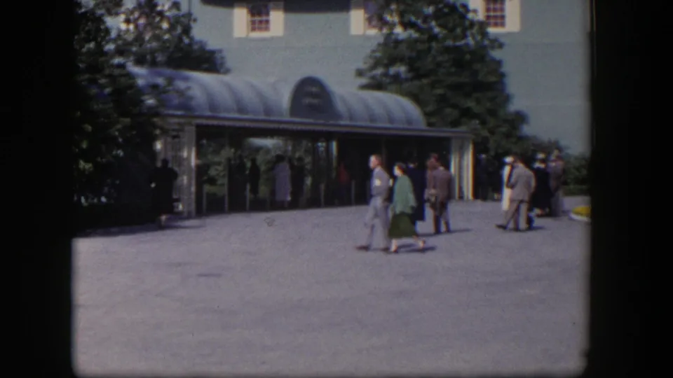 1954 Las Vegas 8mm frame showing adults in mid-century clothing crossing a sunny courtyard before a glass entrance canopy.