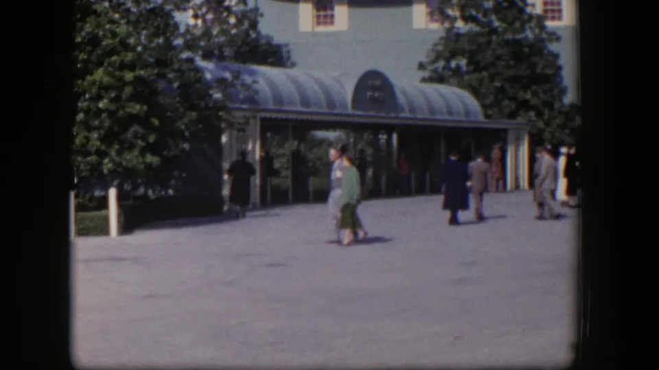 Adults in 1950s attire walk near a landscaped Las Vegas entrance with a curved glass canopy and shaded doorway.