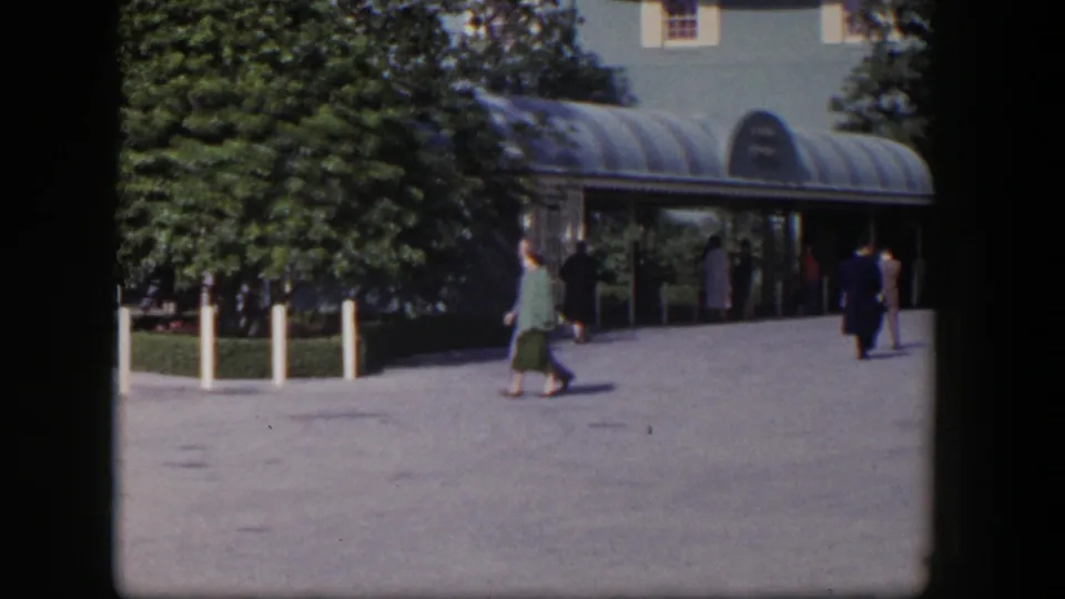 A woman in a green outfit crosses the foreground as adults stand or walk near a glass-canopied Las Vegas entrance.